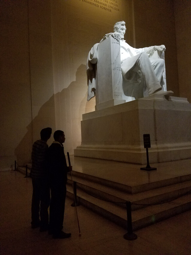During their fall 2016 visit to the Lincoln Memorial, Sushil Adhikari, from Nepal (right), and Nang Attal, from Afghanistan, discovered there was no Audio Description available at the site. So, Attal read the wall text to Adhikari and did his best to describe the surroundings, illustrating a latent need for Audio Description at this precious public resource. The two visitors to Washington, D.C., attended the same conference but did not know each other before this trip. They quickly had become friends, though, and decided to visit the Lincoln Memorial on this evening. In this image, the two are in silhouette, standing next to each other at the base of the statue. The scene is mostly dark, except for Lincoln's illuminated and oversized figure, sitting in a chair, as he stares straight ahead and out toward the National Mall's Reflecting Pond. Adhikari and Attal are on Lincoln's right side, closest to his right hand, as his fingers drape over the arm of his chair. Lincoln's left hand is balled into a fist. Attal is looking up at the statue and describing it. Adhikari, with his cane in his right hand, is listening carefully to Attal and his descriptions. The UniD Project is intended to help visitors, like Adhikari, who are blind or visually impaired, have equivalent experiences to anyone else in this place. UniD-supported descriptions, vetted by park staff, allow more people to participate fully in important societal and cultural discussions, including people who are blind, deafblind, or low-vision. As part of this project's Descriptathon 6, the UniD team is working with the Lincoln Memorial this fall (2020) to audio describe this iconic memorial as well as a dozen other NPS sites around the Washington, D.C., area. When those are complete, they will be uploaded to the free UniD app.